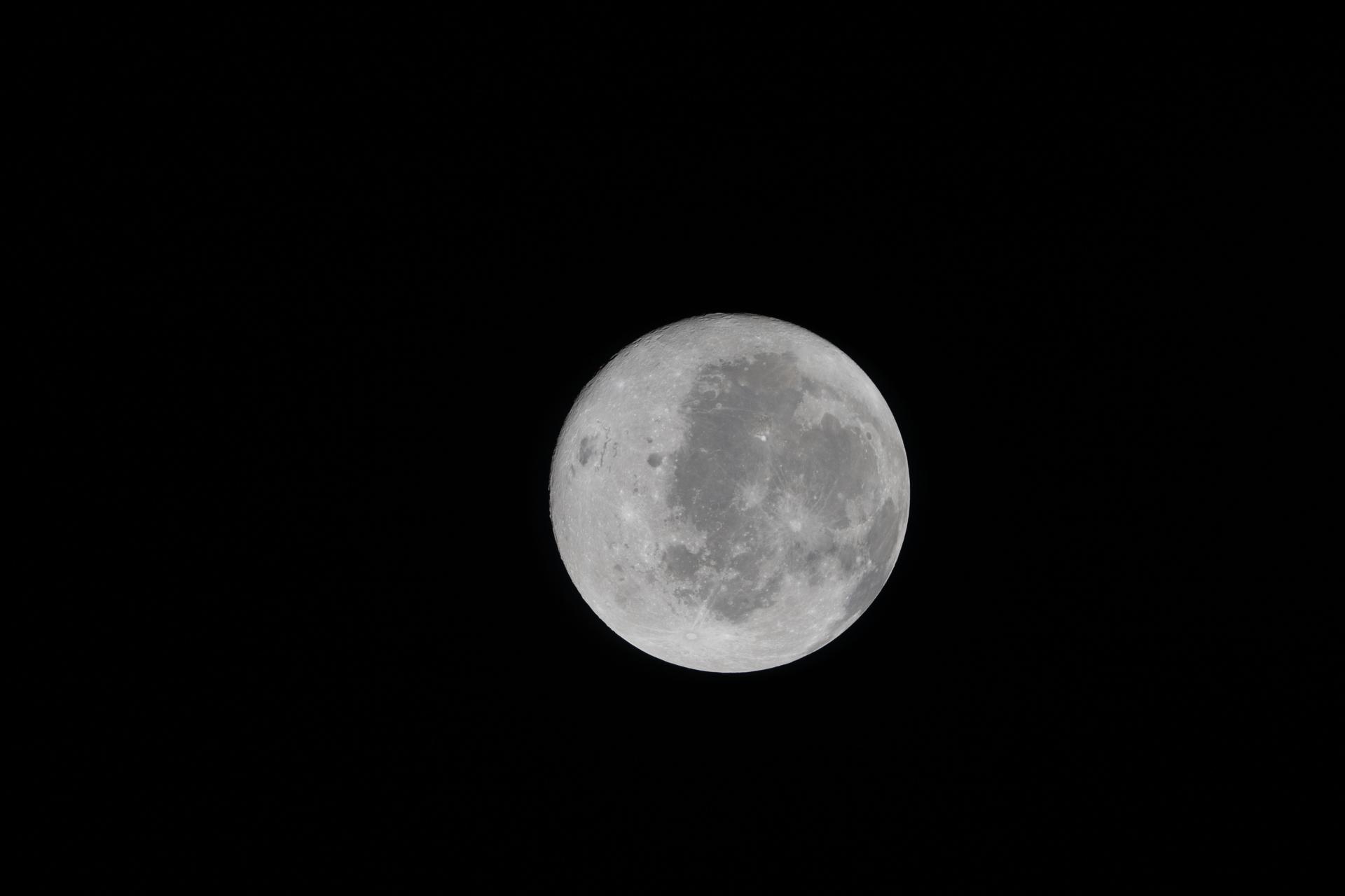 A detailed, high-resolution photograph of the full moon centered against a completely black night sky. The moon appears bright white with various shades of gray, showing its textured surface clearly. Visible are numerous craters, darker lunar maria (large, flat basaltic plains), and lighter highland areas. The moon’s round shape is well-defined, and the contrast between the illuminated surface and the dark sky highlights the moon’s detailed topography. No other objects, stars, or light sources are visible in the image. The overall composition focuses solely on the moon, emphasizing its natural features and surface details.