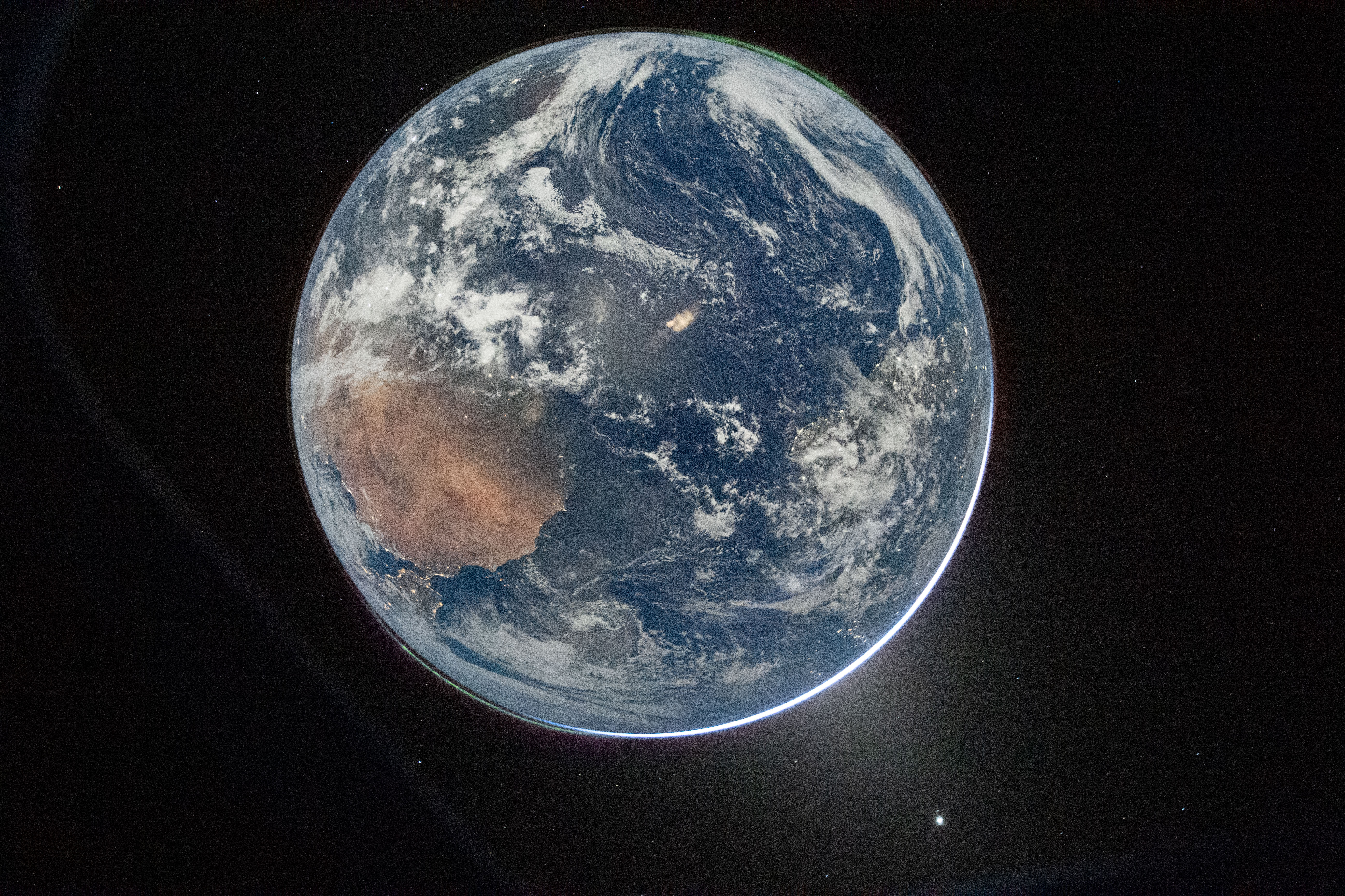 A detailed image of Earth taken from space, showing the planet against a black background dotted with small stars. The view focuses on the Eastern Hemisphere, prominently featuring Australia with its reddish-brown landmass on the left side of the globe. Surrounding Australia are vast expanses of deep blue ocean with swirling white cloud formations scattered across the atmosphere. The curvature of the Earth is clearly visible, with a thin, bright blue atmospheric glow outlining the planet’s edge. Near the bottom right of the image, a bright white star or planet is visible in space. The overall scene captures the beauty and fragility of Earth from a distant vantage point in space.