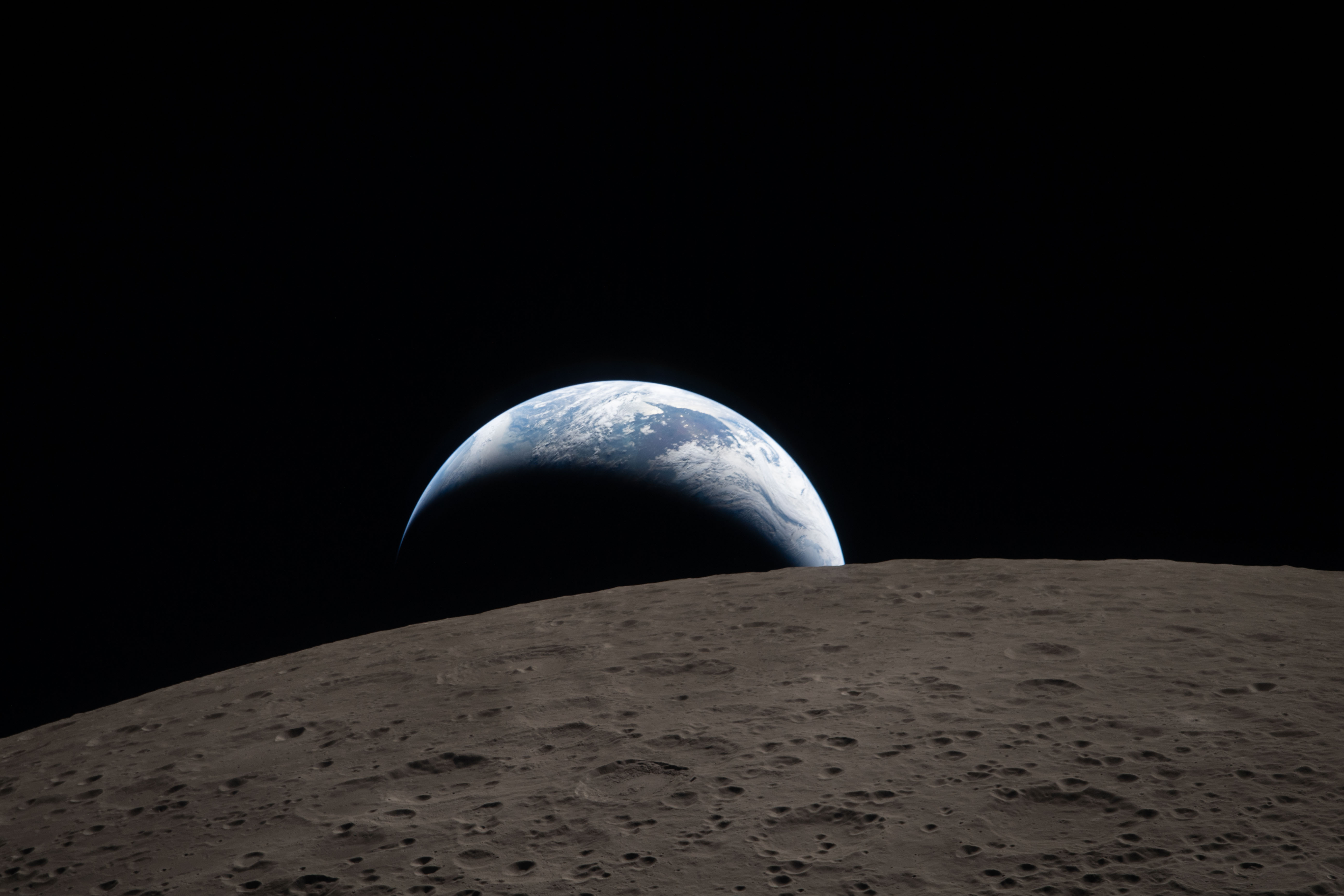 A view from the surface of the Moon showing its gray, cratered terrain in the foreground. The lunar surface is covered with numerous small and medium-sized impact craters, giving it a rough and pockmarked appearance. Beyond the Moon's horizon, the Earth is visible partially illuminated against the blackness of space. The Earth appears as a blue and white crescent with visible cloud formations and oceanic areas, with the shadowed portion blending into the dark background. The image captures the stark contrast between the barren, cratered lunar surface and the vibrant, cloud-covered Earth rising above it. The overall scene conveys a sense of vastness and isolation in space.