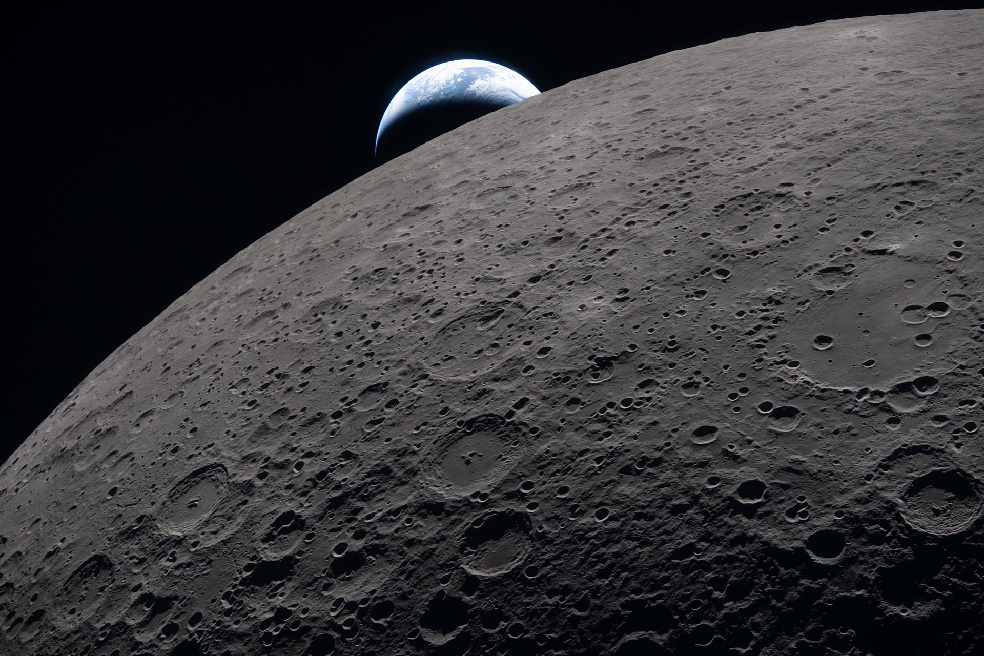 A detailed view of the Moon's surface dominates the foreground, showing a vast expanse covered with numerous craters of varying sizes and depths. The surface appears gray and textured, with shadows accentuating the rugged terrain and crater rims. In the background, partially visible above the Moon's horizon, is the Earth, appearing as a bright, blue and white sphere. The Earth’s surface shows cloud formations and oceanic areas, illuminated by sunlight, contrasting sharply against the blackness of space. The image captures the stark contrast between the barren, cratered lunar surface and the vibrant, life-supporting Earth rising behind it. The overall scene conveys a sense of vastness and isolation in space.