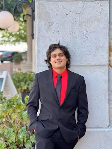 A young man celebrating his 21st birthday stands confidently in a black suit, red shirt, and black tie, leaning against a light gray stone wall. He smiles softly with his hands in his pockets, and decorative lights and greenery appear in the background.