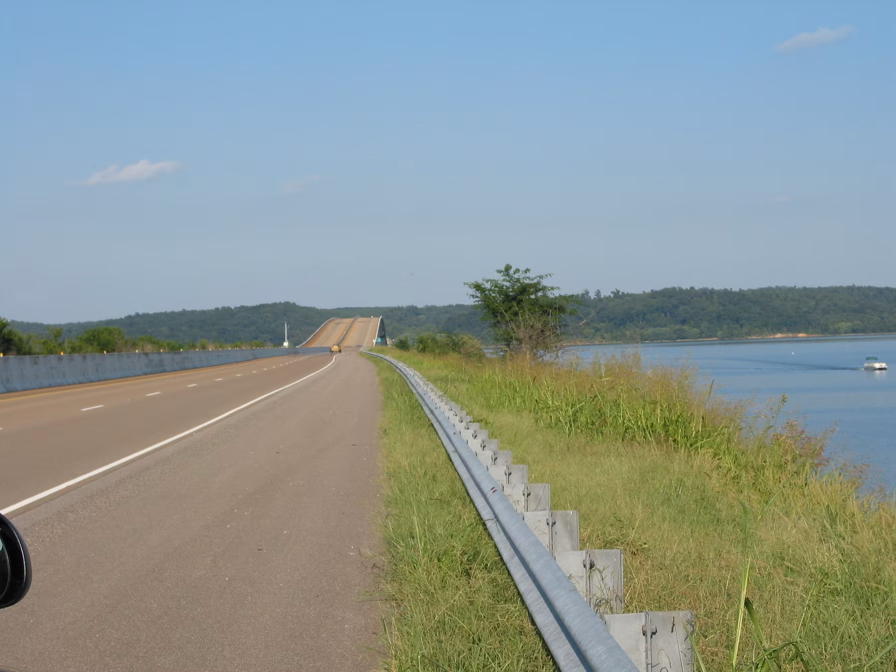 A long, empty road runs parallel with a guardrail next to a body of water, bordered by grass and trees under a clear blue sky.