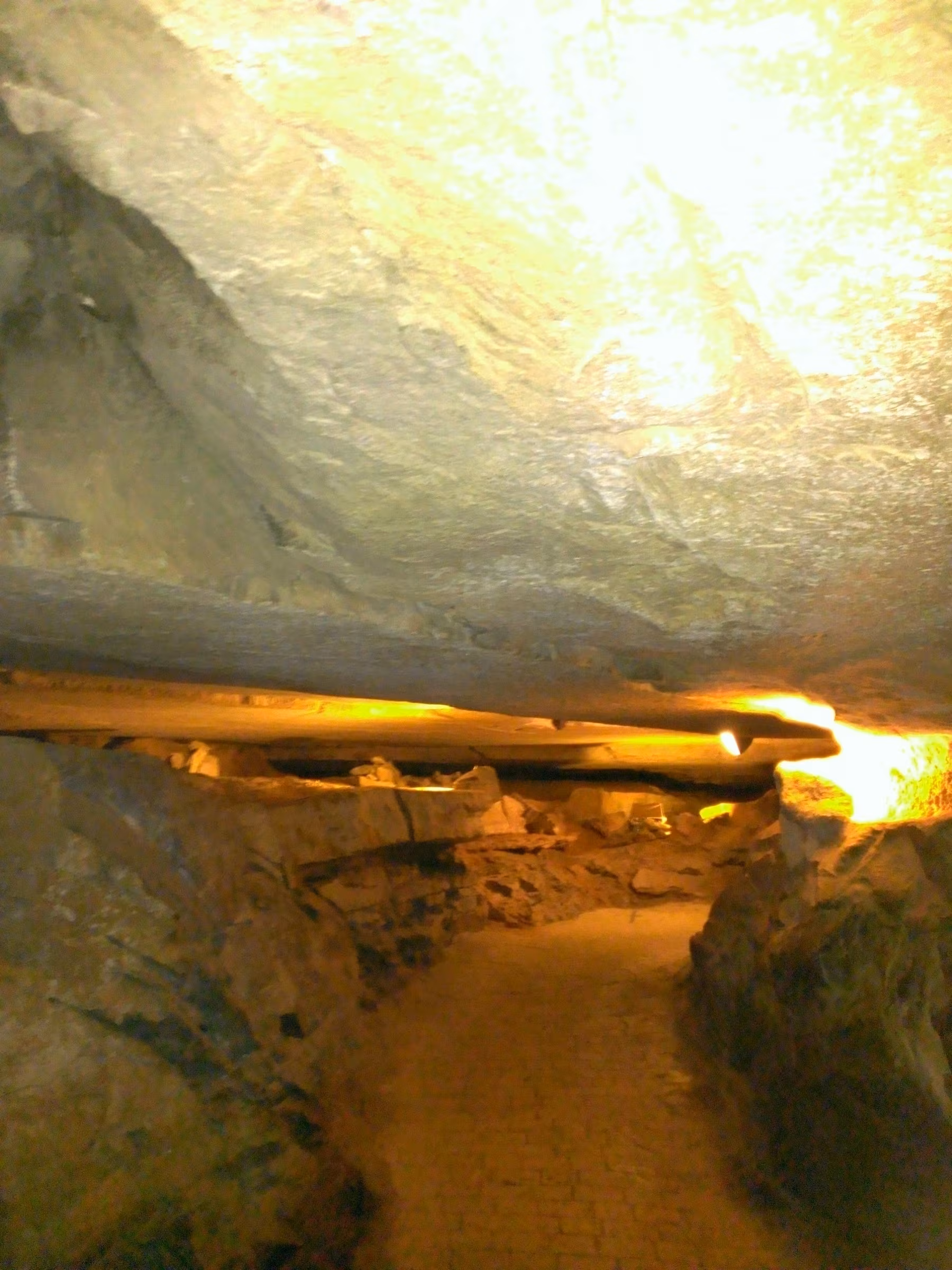 A narrow cave pathway is illuminated by warm overhead lighting, highlighting the rocky ceiling and walls.