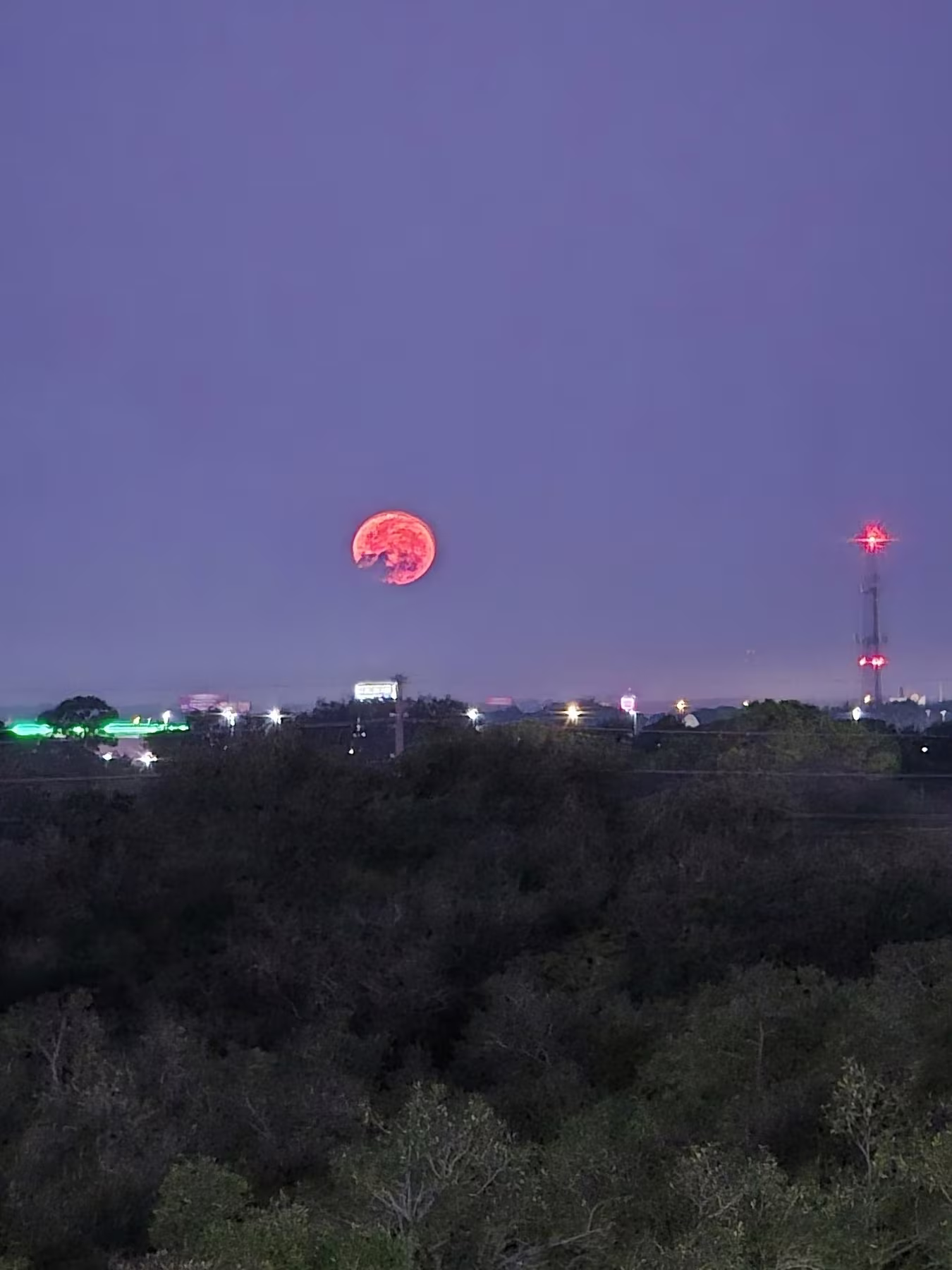 A glowing red moon rises over a landscape with trees and distant city lights against a dusky sky.