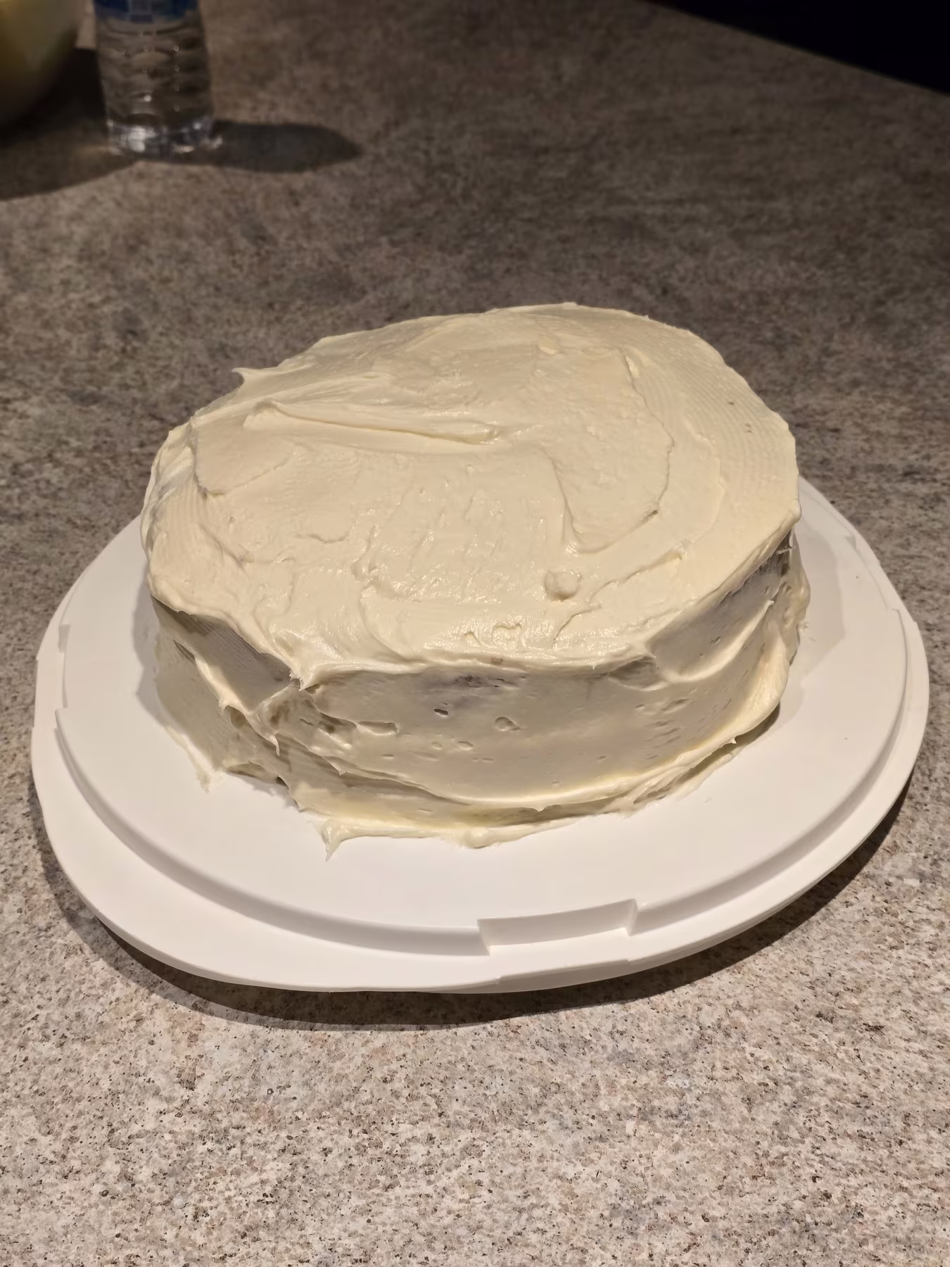 A round cake covered with white frosting is placed on a white platter on a countertop.