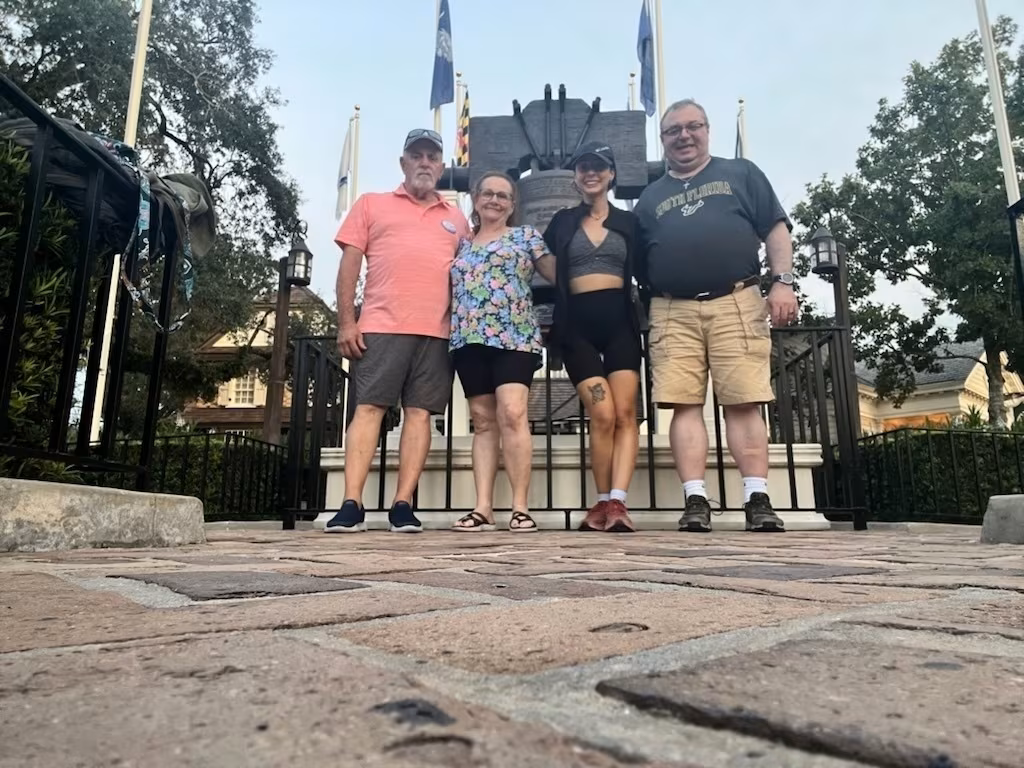 Four people are standing together on a cobblestone path in what appears to be a park or recreational area, with flags and structures in the background.