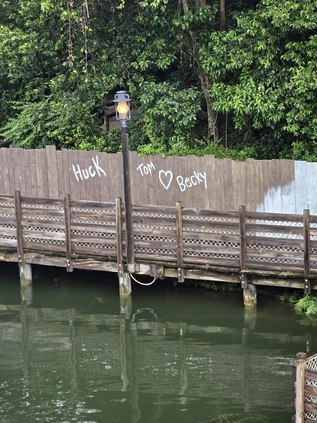 A wooden walkway over water leads to a graffiti-marked fence with lush greenery in the background.