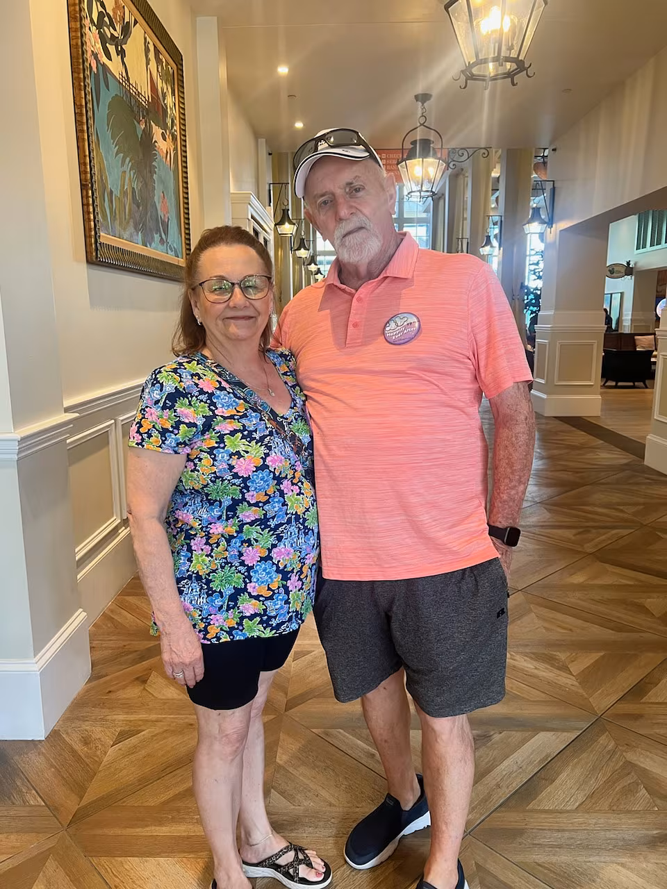 A smiling older couple stands arm-in-arm in a beautifully decorated hallway at Disney World, with warm lighting, wooden floors, and hanging lanterns. The woman wears a colorful floral top and black shorts, and the man wears a coral polo shirt, dark shorts, a white cap, and a celebratory button. The photo was taken during their anniversary celebration trip