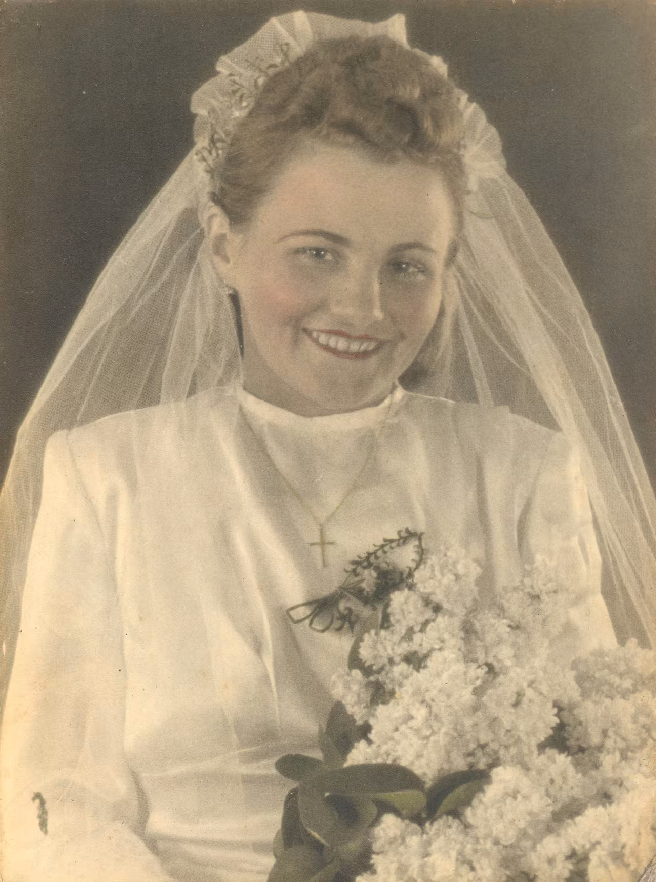 A vintage portrait of a smiling bride in her wedding attire. She wears a white gown with a high collar and a delicate cross necklace. Her hair is styled in elegant curls topped with a floral headpiece and a flowing veil. She holds a large bouquet of white flowers, adding a classic touch to her joyful expression.