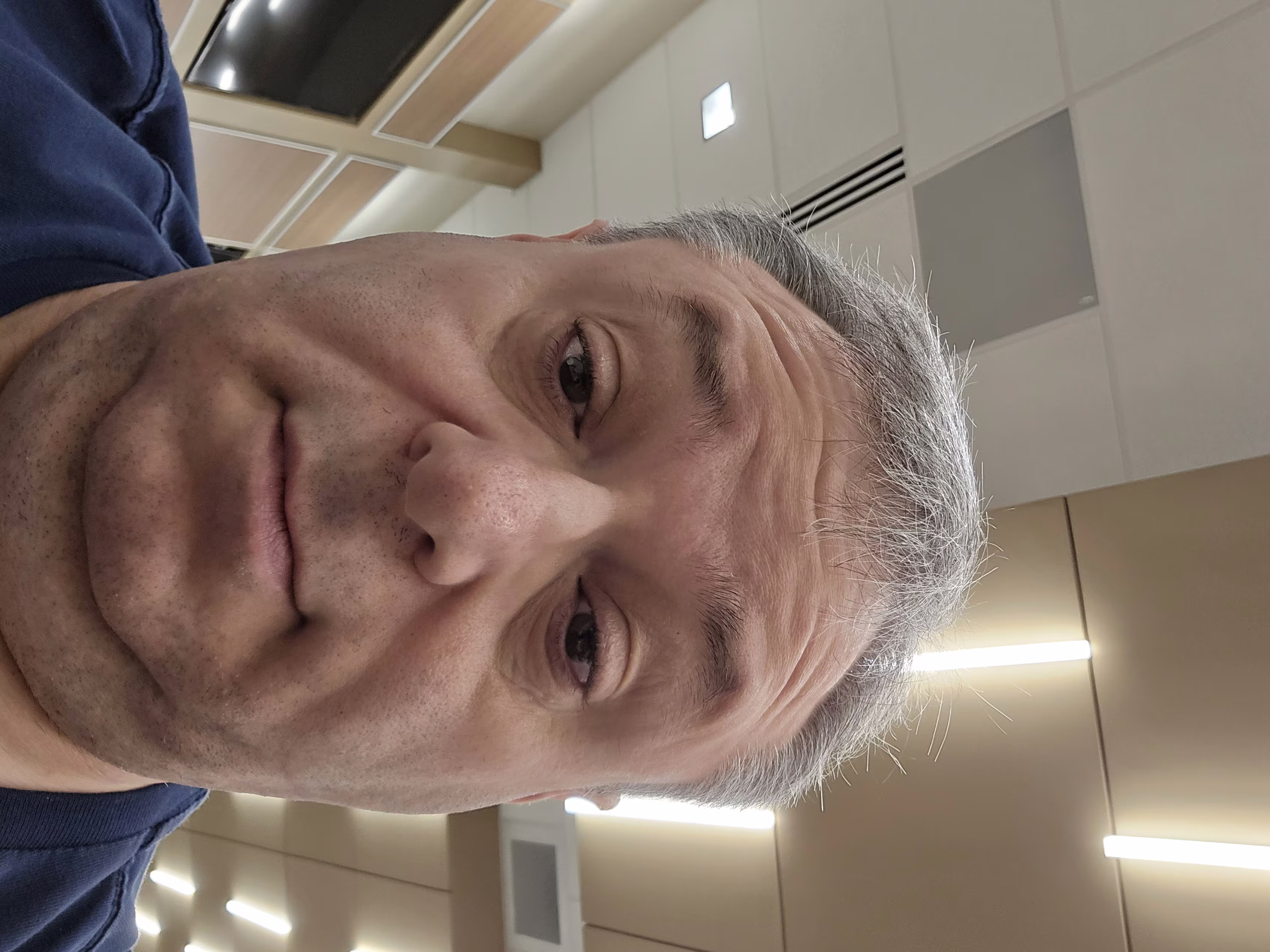 Close-up image of a middle-aged man with light skin and short, graying hair. The photo is taken from a low angle, showing the upper half of his face and part of his neck and shoulders. His eyes are partially open, and he has a neutral expression. The man is wearing a dark blue shirt. The background shows a ceiling with beige and white panels, fluorescent lights, and part of a wall with a mounted TV screen and wooden paneling. The lighting is bright and evenly distributed. The image captures a candid, casual moment indoors.