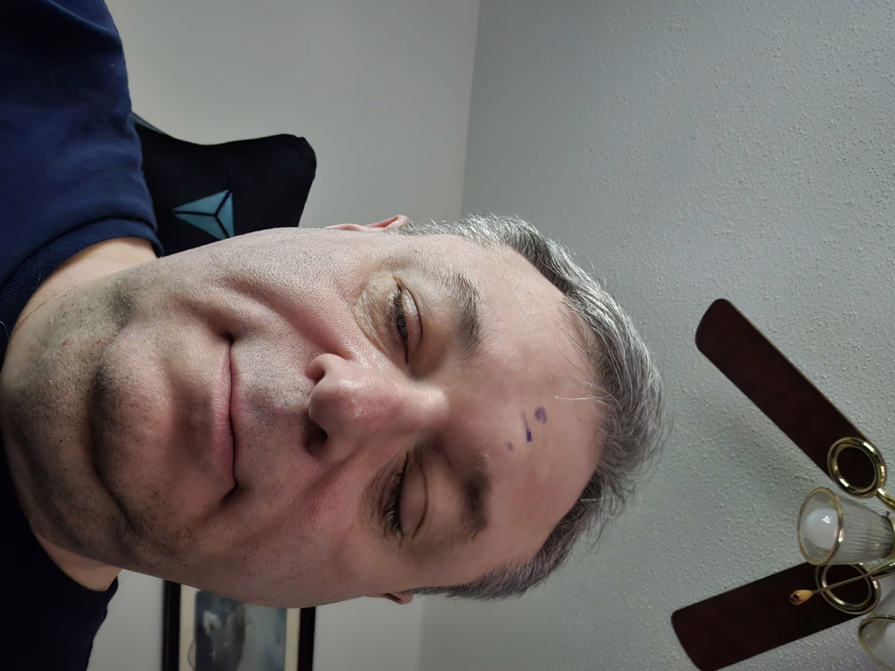 A close-up selfie of a middle-aged man with light skin and short, graying hair. His eyes are partially closed, and he has a neutral expression with a slight smile. On his forehead, there are three small purple marks, possibly made with a pen or marker. He is wearing a dark blue shirt. Behind him, part of a black chair with a blue geometric design is visible. The background shows a white textured ceiling with a ceiling fan that has dark brown blades and brass fixtures, including a light with a frosted glass cover. A framed picture is partially visible on the wall to the right. The lighting is natural and soft.