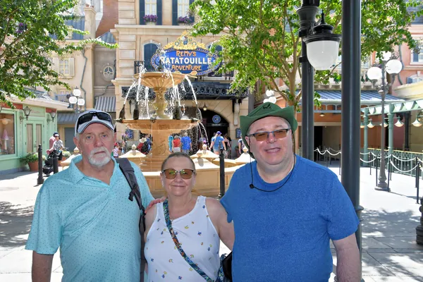 Three people in front of the France pavilion fountain at Epcot with Remy's Ratatouille Adventure behind them