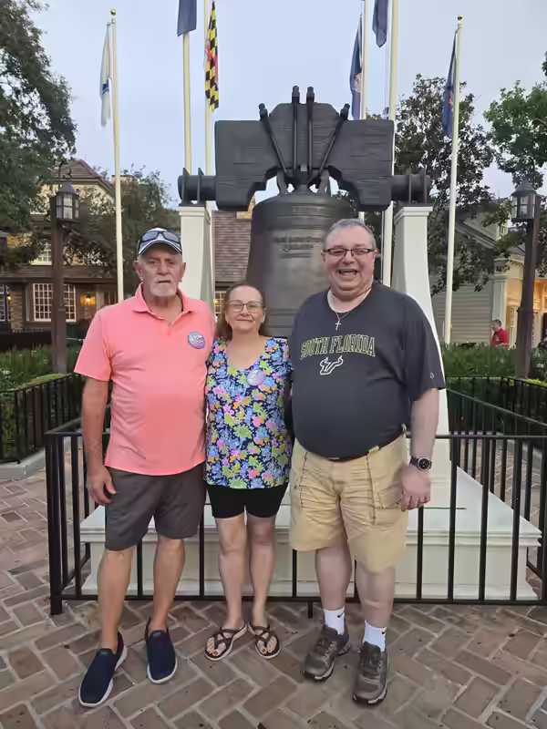 Three people posing in front of the Liberty Bell replica in Liberty Square at Magic Kingdom, with American flags and colonial brick architecture in the background