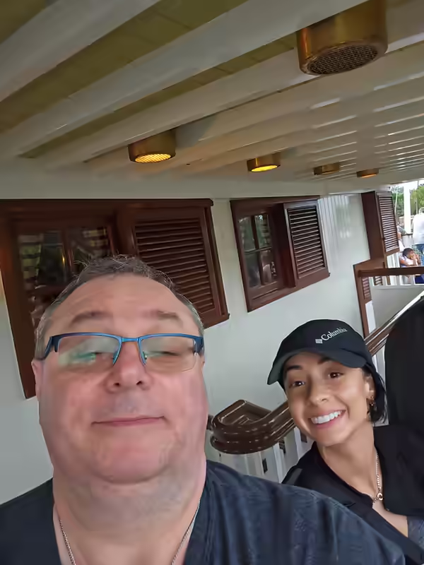 Man with blue glasses and young woman in a Columbia cap taking a selfie aboard the Liberty Belle Riverboat, with the boat's white Victorian woodwork and brass wall sconces visible behind them