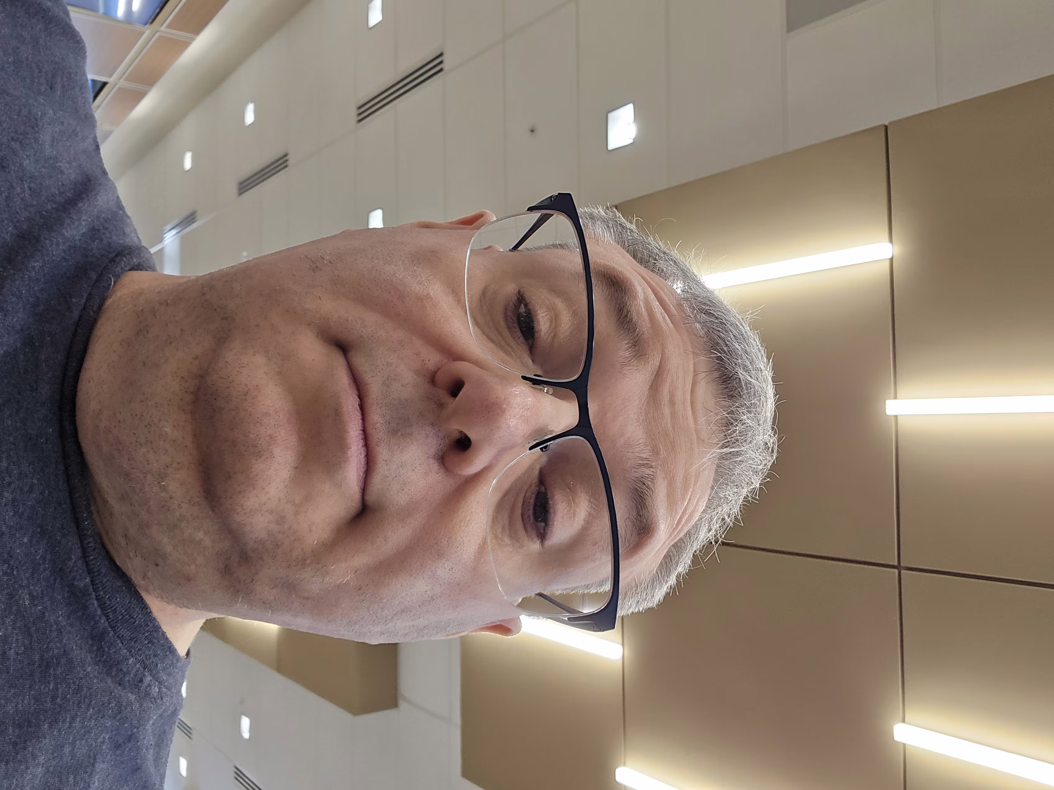 Close-up selfie of a middle-aged man with short gray hair and light skin, wearing rectangular black-framed glasses. The photo is taken from a low angle, capturing the lower half of his face prominently, including his nose, mouth, and chin, with a neutral expression. He is wearing a dark blue shirt. The background shows an indoor setting with a ceiling featuring beige panels and long, bright fluorescent lights arranged in parallel lines. Additional ceiling elements include white square tiles and air vents. The lighting is bright and evenly distributed.