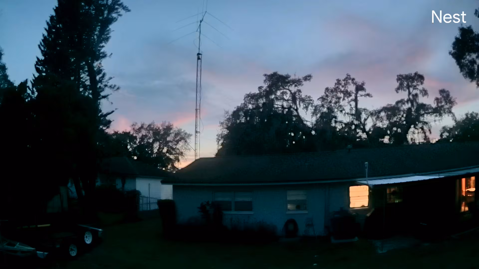 A backyard view just after sunset showing a Tempest Weather Station mounted high above a house, silhouetted against a blue, pink, and purple evening sky.